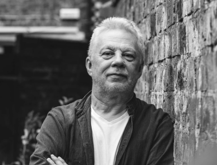 Black-and-white portrait of a person standing beside a textured brick wall, facing the camera with arms crossed. The person has short light-colored hair and is wearing a light T-shirt under an open dark jacket. The background is softly blurred, drawing attention to the subject and the brick wall